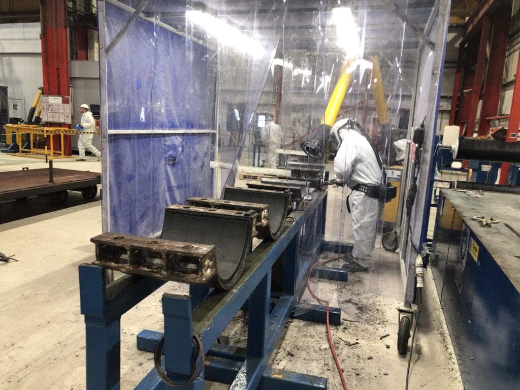 Technician in PPE applying coating to steel half-shell clamps inside a protective booth at LJF Custom Coatings workshop