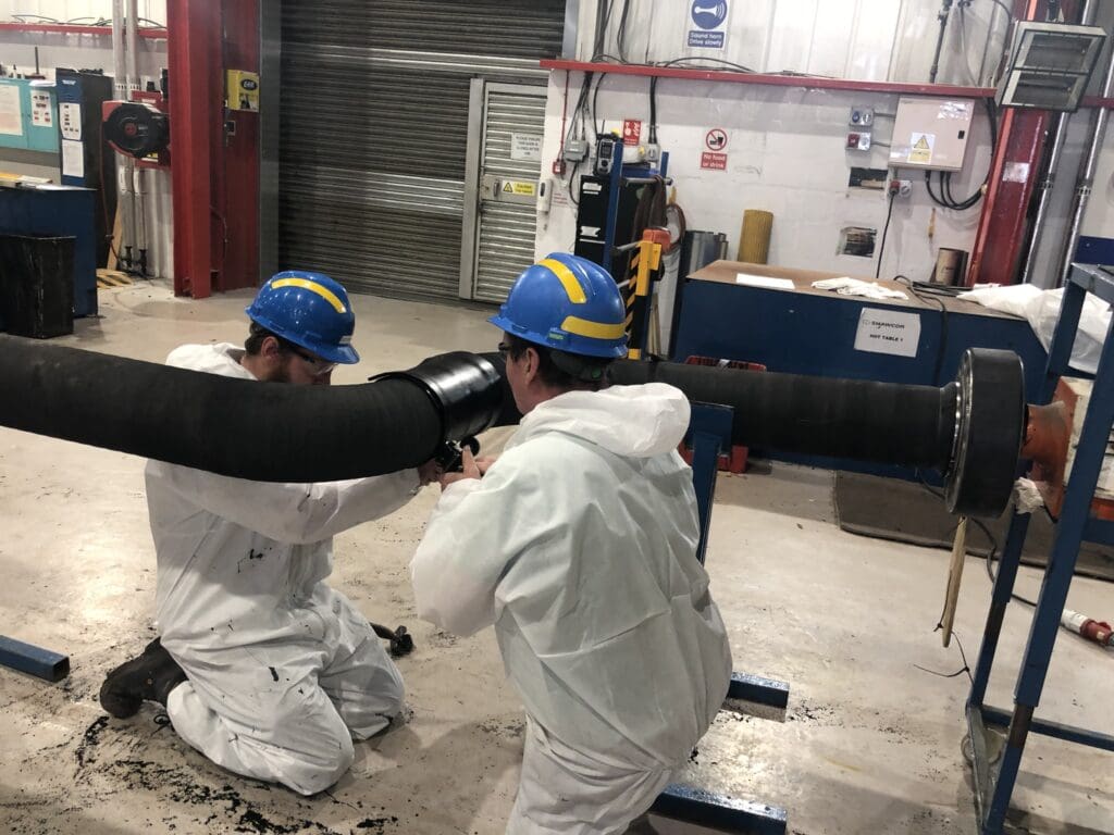 Two LJF Custom Coatings technicians in PPE installing and sealing a Neoprene rubber joint on a black-coated subsea pipeline section