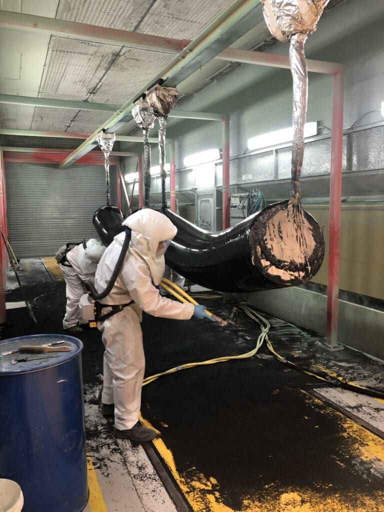 LJF Custom Coatings technician in PPE applying anti-corrosion coating to suspended pipe section inside spray booth